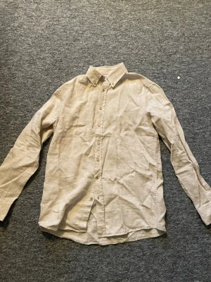 Beige linneskjorta med krage - Snygg beige skjorta i linne med klassisk krage och knäppning framtill. Skjortan har lång ärm och en avslappnad passform, perfekt för en stilren och enkel look.