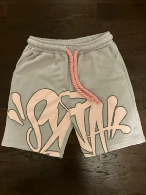 Grå shorts med rosa graffiti-tryck  - Säljer ett par grå syna world shorts med stort rosa tryck framtill och breda rosa snören i midjan. Shortsen har elastisk midja och djupa sidofickor, kvaliteten är riktigt hög, perfekta för en avslappnad streetwear-look.