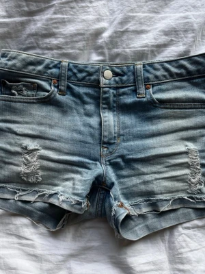 Ljusblå slitna jeansshorts - Säljer ett par ljusblå jeansshorts med slitna detaljer och fransig kant. Shortsen har klassisk femficksdesign, knappgylf och en relaxed passform. Perfekta för varma dagar och en avslappnad stil.