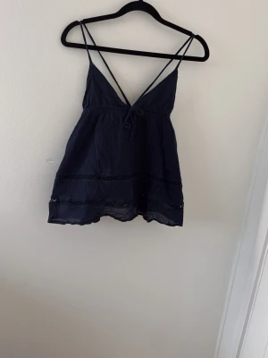 Mörkblå linnetopp med spetsdetaljer - Snygg mörkblå linnetopp med tunna axelband som korsas i ryggen. Toppen har v-ringning och dekorativa spetsdetaljer längs nederkanten och mitten. Perfekt till sommaren och enkel att matcha med jeans eller kjol.
