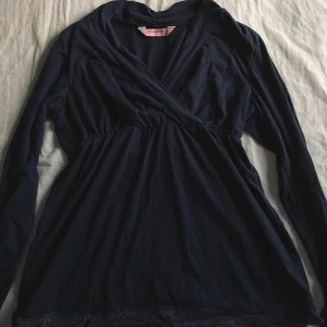 Mörkblå topp från Hunkemöller - Mörkblå topp eller nattlinne från Hunkemöller med långa ärmar och omlott-ringning. Tillverkat i mjukt tyg och har en elastisk midja samt spetsdetalj längst ner. Perfekt för mysiga kvällar hemma.