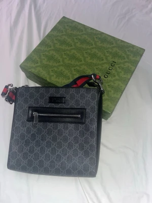 Gucci GG Supreme axelväska - Snygg axelväska från Gucci i GG Supreme-mönstrad canvas med svarta läderdetaljer. Väskan har en framficka med dragkedja och justerbar axelrem i rött och blått. Kommer med originalbox, dustbag och äkthetskort.