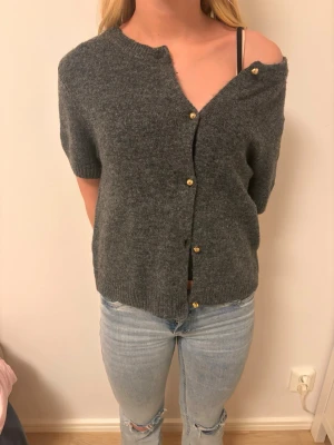 Mörkgrå kofta med kort ärm - Mysig mörkgrå kofta med korta ärmar och guldfärgade knappar. Koftan har en rak passform och ribbade kanter vid ärmslut och nederkant. Perfekt att slänga över en topp för en avslappnad look.