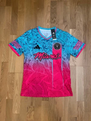 Adidas Inter Miami FC tröja sopran - Säljer en färgstark Inter Miami FC tröja från Adidas med blått, rosa och lila mönster. Tröjan har korta ärmar, V-ringning och trycket 'Miami' på bröstet samt klubbmärke. Tillverkad i lätt och ventilerande polyester, perfekt för träning eller match.