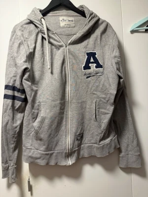Grå zip hoodie Iconic Sport - Grå hoodie med dragkedja från Iconic Sport. Stor blå bokstav 'A' och text på bröstet, två mörkblå ränder på ena ärmen och fickor framtill. Huva med vit dragsko. Mjuk bomullsmix som känns skön mot huden. Perfekt för en avslappnad streetstyle.