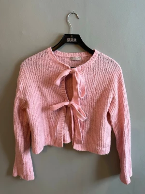 Rosa stickad kofta med rosettknäppning - Söt rosa stickad kofta med två rosetter framtill som knäppning. Koftan är croppad med långa ärmar och har en mjuk, ribbstickad struktur. Perfekt för lager-på-lager och ger en gullig touch till din outfit. Pris förslag skickas gärna