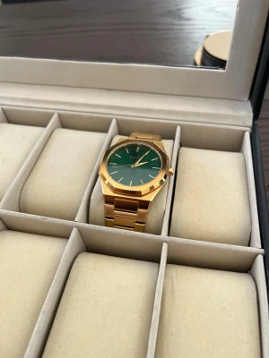 Ekstrand Guldfärgad klocka med grön urtavla - Stilren klocka med guldfärgad länk i metall och en djupgrön urtavla. Klockan har minimalistisk design med tunna visare och index, samt en robust men elegant känsla. Perfekt accessoar för dig som vill sticka ut med klassisk stil.