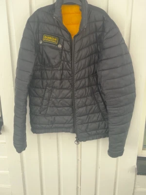Svart dunjacka från Barbour - Snygg svart dunjacka från Barbour International med gul insida och patch på bröstet. Jackan har quiltat mönster, dragkedja framtill och praktiska fickor. Perfekt för kalla vinterdagar och ger en cool streetwear-look.