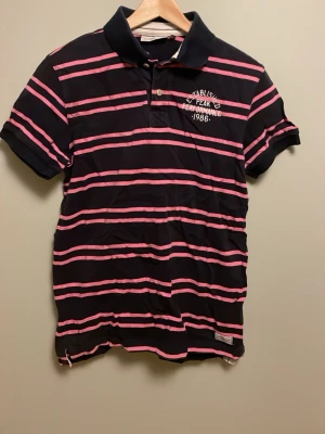 Vintage randig pikétröja Peak Performance - Mörkblå pikétröja från Peak Performance med rosa ränder och broderad logga på bröstet. Klassisk krage, korta ärmar och knappar framtill. Tillverkad i bomull för en skön känsla och sportig look.