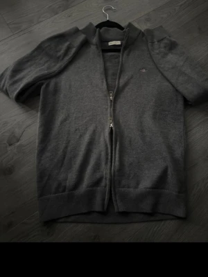 Mörkgrå ziptröja från Gant - Mörkgrå ziptröja från Gant  med diskret logga på bröstet. Tröjan har lång ärm, ribbade muddar och hög krage. Tillverkad i mjukt stickat material som ger en clean och stilren look. Perfekt för lager på lager och enkel att matcha.