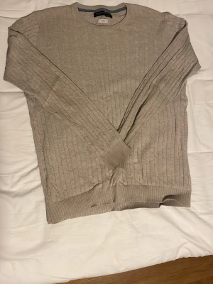 Beige stickad tröja från Jack and Jones  - Beige stickad tröja från Jack and Jones med rund halsringning och ribbade muddar. Tröjan har ett diskret kabelstickat mönster och är tillverkad i ett mjukt material som känns skönt mot huden. Perfekt för lager på lager och enkel att matcha. Tröjan är i barn storlek large och passar dig som är ca 175