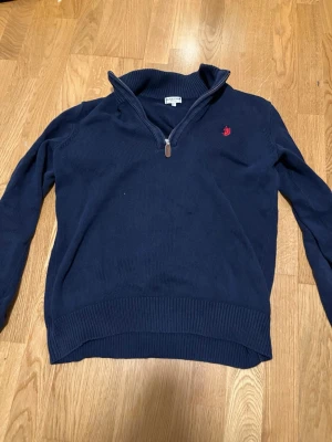 Mörkblå stickad tröja U.S. Polo Assn. - Mörkblå stickad tröja från U.S. Polo Assn. med halv dragkedja och röd broderad logga på bröstet. Tröjan har ribbstickade muddar och krage, perfekt för lager-på-lager-stil. Klassisk och stilren design.