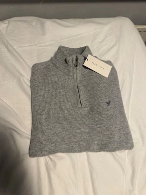 Pier One stickat tröja  - Snygg ljusgrå stickad tröja från Pier One med halv dragkedja och hög krage. Tröjan har ett diskret broderat motiv på bröstet och är ribbstickad för extra komfort. Perfekt för lager på lager och enkel att matcha.