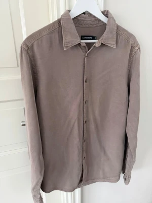 Beige skjorta från J.Lindeberg - Stilren beige skjorta ” slim LS comfort tencel shirt”från J.Lindeberg i mjukt tencel-material. Skjortan har klassisk krage, knäppning framtill och långa ärmar. Passformen är slim och den har en enkel, tidlös design som passar till många olika looks.