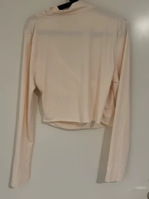 Ljusbeige omlottopp med drapering - Trendig ljusbeige omlottopp med draperad front och långa ärmar. Toppen har croppad passform och är tillverkad i mjukt material som känns skönt mot huden. Perfekt för dig som gillar stilrena och moderna plagg.