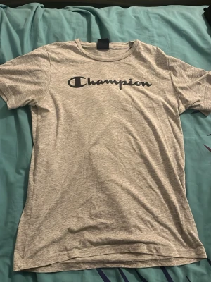 Grå Champion t-shirt med logga - Snygg grå t-shirt från Champion med stor svart logga över bröstet. Klassisk passform med rund hals och korta ärmar. Perfekt för en avslappnad och sportig stil. Tillverkad i mjuk bomull för extra komfort.