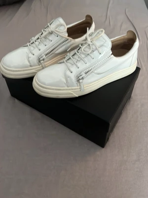 Vita Giuseppe Zanotti skor - Snygga vita sneakers från Giuseppe Zanotti i glansigt lackmaterial. Skorna är i väldigt bra skick dem har blivit använda men väldigt fräscha. Perfekta för dig som vill sticka ut med en clean och modern look. Fler frågor är de bara att skriva så svarar jag. Storlek 43.