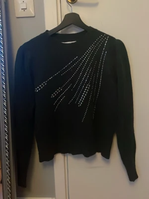 Svart stickad tröja med strassdetaljer - Snygg svart stickad tröja med långa ärmar och coola strassdetaljer i ett mönster över ena axeln och bröstet. Tröjan har rund halsringning och är lite croppad i modellen. Perfekt för dig som vill ha något extra till din outfit. Från only