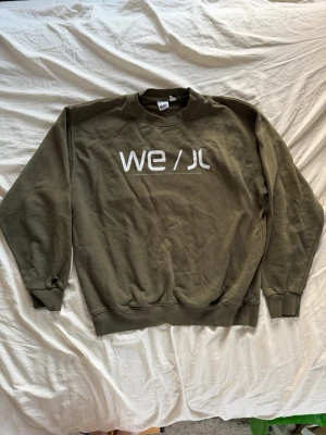 Olivgrön sweatshirt från WE/ JL - Olivgrön sweatshirt med rund hals och tryckt vit WE/ JL-logga på bröstet. Tröjan har ribbade muddar vid ärmslut och nederkant, samt en avslappnad passform. Perfekt för en casual och stilren look. Tyvärr finns det ett hål vid höger hand (se bild). Trycket är inte i sitt bästa skick (se bild)