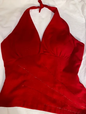 Röd glittrig halterneck topp - Snygg röd halterneck topp med glittriga detaljer och djup v-ringning. Toppen knyts i nacken och har dragkedja i ryggen. Passar dig som vill ha en statement piece, storlek S/M. Säljer för den inte kommer till användning 