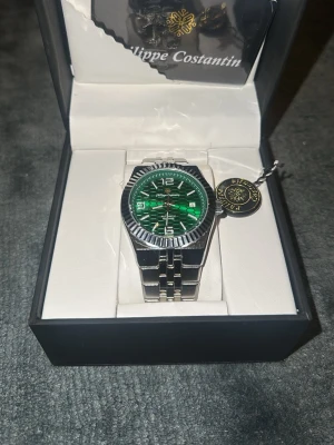 Grön Philippe Costantin klocka - Snygg klocka från Philippe Costantin med ett grönt, mönstrat urverk och silverfärgad länk i metall. Klockan har en unik urtavla med geometriskt mönster och tydliga siffror. Perfekt accessoar för dig som vill sticka ut med stilren design.