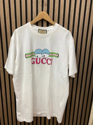 Vit t-shirt med tryck - Vit t-shirt  med färgglatt tryck framtill som visar två blå ögon och gröna, rosa och svarta detaljer. T-shirten har klassisk passform, rund halsringning och är tillverkad i mjuk bomull. Perfekt för dig som vill sticka ut med en unik designerstil.