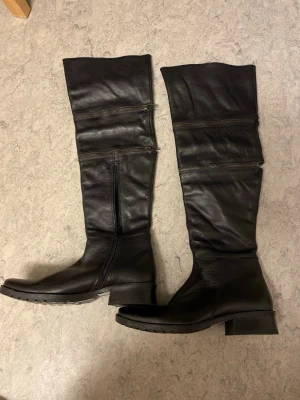 Svarta knähöga läderstövlar med dragkedjor - Säljer ett par svarta knähöga stövlar i äkta läder med grov sula och dekorativa dragkedjor längs skaftet. Klassisk design med låg klack och rundad tå, perfekta till höstens och vinterns outfits.