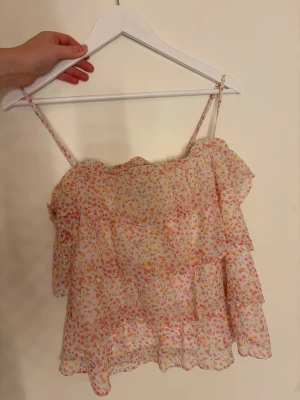 Blommig volangtop från Vero Moda - Supersöt blommig topp från Vero Moda med tunna axelband och volangdetaljer. Toppen är vit med små blommor i rosa, orange och gul, och har en luftig, lätt känsla – perfekt för varma dagar.