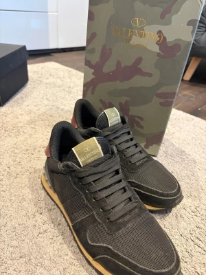 Svarta Valentino sneakers rockrunner  - Snygga svarta sneakers från Valentino med coola nitar på hälen och detaljer i mocka och mesh. Skorna har snörning och en beige sula som ger en schysst kontrast. Perfekta för dig som vill ha en stilren men edgy look.