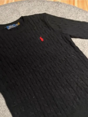 Svart kabelstickad tröja Polo Ralph Lauren - Kabelstickad svart tröja från Polo Ralph Lauren med rund halsringning och röd broderad logga på bröstet. Tröjan har långa ärmar och ribbade muddar vid ärmslut och nederkant. Klassisk och stilren design i mjukt material. Storlek XXL men passar bra på mig som är en L vanligtvis
