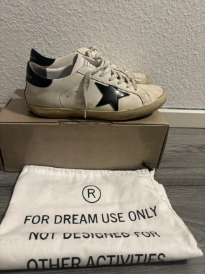 Golden Goose vita sneakers med svart stjärna - Golden Goose sneakers i vitt läder med svart stjärndetalj på sidan och svart hälparti. Klassisk låg modell med vita skosnören och beige sula. Snygg streetstil och ikonisk design som sticker ut.