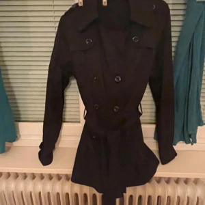 Svart trenchcoat från Vila, storlek L - Stilren svart trenchcoat från Vila med dubbelknäppning, klassisk krage och bälte i midjan. Jackan har långa ärmar och dekorativa axelklaffar. Perfekt för dig som vill ha en tidlös och snygg look.