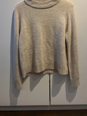 Beige stickad tröja från Wera - Mysig beige stickad tröja från Wera med hög, ribbad krage och långa ärmar. Tröjan har ribbade muddar vid ärmslut och nederkant, perfekt för lager-på-lager. Enkel och stilren design som passar till det mesta.