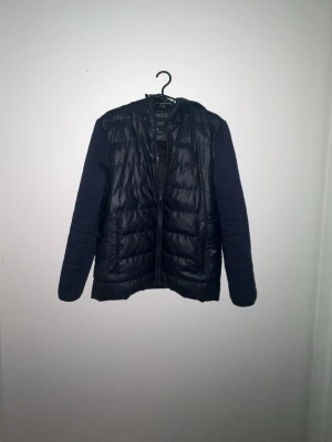 Mörkblå dunjacka med huva - Mörkblå svart dunjacka med quiltat mönster och huva. Jackan har dragkedja framtill, två sidofickor och mjuka ärmar i tyg. Perfekt för kalla dagar och enkel att matcha med olika outfits. Litet märke som finns på ena armen inget stort