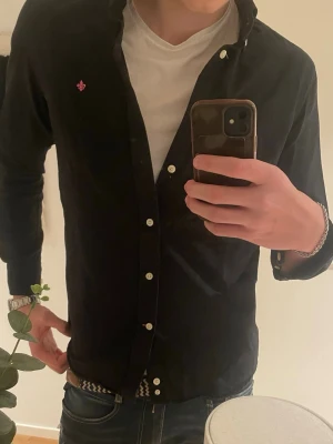 Svart skjorta från Morris Stockholm - Säljer en svart skjorta från Morris Stockholm i storlek M. Skjortan har klassisk krage, vita knappar och en liten broderad logga i rött på bröstet. Tillverkad i mjuk bomull och passar perfekt till jeans eller chinos.