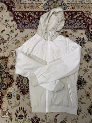 Nike vit och beige vindjacka med huva - Nike vindjacka i vitt och beige med dragkedja framtill och två sidofickor. Jackan har en justerbar huva och ribbade muddar vid ärmslut och nederkant. Tillverkad i lätt, vindtätt material som passar perfekt för blåsiga dagar.