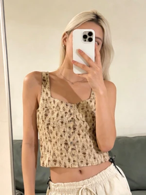 Beige blommig linnetopp med knappar - Superfin croppad linnetopp i beige med små blommigt mönster och knappar framtill. Toppen har breda axelband och fyrkantig ringning, perfekt för varma dagar. Lätt och luftig känsla, passar snyggt till både kjol och byxor.