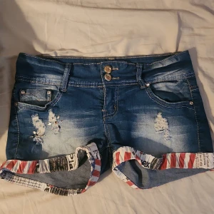 Lågmidjade jeansshorts - Snygga blå lågmidjade jeansshorts med slitna detaljer och dubbla knappar framtill. Shortsen har uppvikta benslut med coolt tryck i rött, vitt och svart, samt klassiska fickor både fram och bak. Perfekta för en chill sommardag. Måtten syns i bild! Priset kan diskuteras. Knapparna känns lite lösa men dem var så när jag köpte shortsen (det är alltså meningen antar jag) och det känns INTE som om de ska ramla av eller något. Köpta på vinted. De är stretchiga! 38cm rakt över midjan 