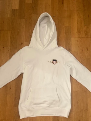 Vit hoodie från GANT med logga - Snygg vit hoodie från GANT med klassisk huva och känguruficka. På bröstet finns GANT:s logga och ett sköldmärke i rött, vitt och blått. Tillverkad i mjukt bomullsmaterial som är skönt att ha på sig. Perfekt för en avslappnad och stilren look.