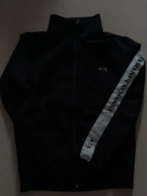 Svart hoodie från Armani Exchange - Snygg svart hoodie från Armani Exchange med dragkedja framtill och vit diagonal detalj med texten 'Armani Exchange' på ena ärmen. Klassisk passform och hög krage. Perfekt för en trendig och sportig look.