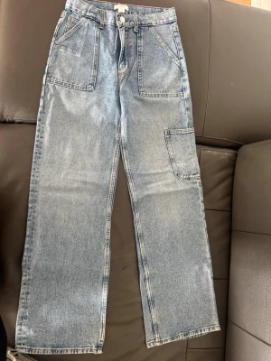 Gina tricot jeans - Säljer ett par ljusblå jeans med raka ben och stora, markerade fickor fram och bak. Jeansen har även en extra sidoficka på benet och är tillverkade i klassisk denim. Perfekta för en avslappnad och trendig look.