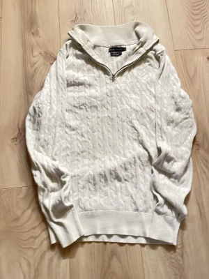 Vit stickad tröja med halvzip SCW - Vit stickad tröja från S.C.W. med halvzip och hög krage. Tröjan har ett klassiskt kabelstickat mönster och är tillverkad av återvunnet material. Perfekt för en stilren och avslappnad look.
