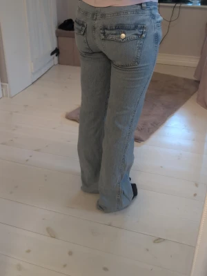 Ljusblå bootcut jeans med fickor - Säljer ett par ljusblå bootcut jeans med klassisk femficksdesign och snygga guldknappar på bakfickorna. Jeansen har en rak passform som är lite utsvängd nedtill och är tillverkade i denim. Perfekta för dig som gillar en avslappnad och trendig look.
