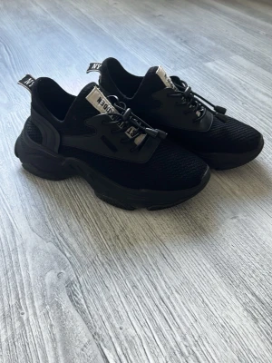Svarta sneakers från Steve Madden  - Stilrena svarta sneakers från Steve Madden  med chunky sula och mesh-ovandel. Skorna har elastiska snören med snabblås och detaljer i svart plast. Märkets logga syns på plösen och hälen. Perfekta för dig som gillar en sportig och modern look.