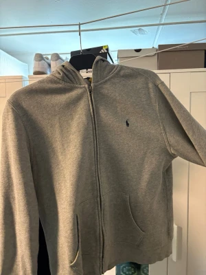 Grå hoodie från Polo Ralph Lauren - Snygg grå zip-hoodie från Polo Ralph Lauren med klassisk huva och fickor framtill. Dragkedja hela vägen och den ikoniska lilla loggan broderad på bröstet. Perfekt för chill dagar eller när du vill ha en clean och enkel look.