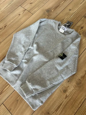 Grå sweatshirt från Stone Island - Säljer en stilren grå sweatshirt från Stone Island med klassisk rund halsringning och ribbade muddar. Tröjan har den ikoniska Stone Island-loggan på vänster ärm och är tillverkad i mjuk bomull. Perfekt för en clean och avslappnad look.
