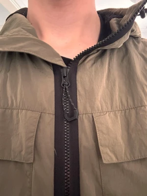 Olivgrön vindjacka från Hackett - Snygg olivgrön vindjacka med dragkedja framtill och två stora bröstfickor. Jackan har huva och justerbara ärmslut med kardborre. Tillverkad i lätt, vindtätt material som passar perfekt för blåsiga dagar. Från Hackett. 