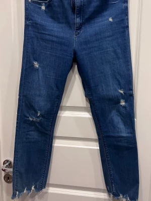 Blå slitna jeans med rå kant från Zara - Säljer ett par blå jeans med slitningar och råa, fransiga benslut. Jeansen har klassisk femficksmodell och är tillverkade i denim med stretch. Perfekta för en avslappnad och trendig look.