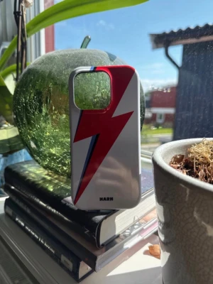 IPhone 16 pro skal från NARN - Snyggt mobilskal i hårdplast från NARM med vit bas och en röd blixt med blå detalj på baksidan. Skalet har svart insida och passar perfekt för dig som vill sticka ut med din mobil. Levereras med en beige tygpåse med logga. 