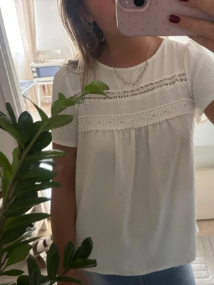 Vit blus med spetsdetaljer - Söt vit blus med korta ärmar och fina spetsdetaljer över bröstet. Blusen har rund halsringning och ett lätt, luftigt tyg med diskret struktur. Perfekt till jeans eller kjol för en fräsch look. Stor i storleken men passar S-L